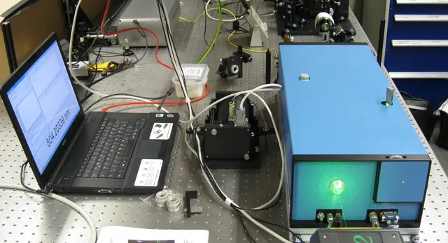 A blue laser housing connected by cables to instruments and a laptop on an optical table.