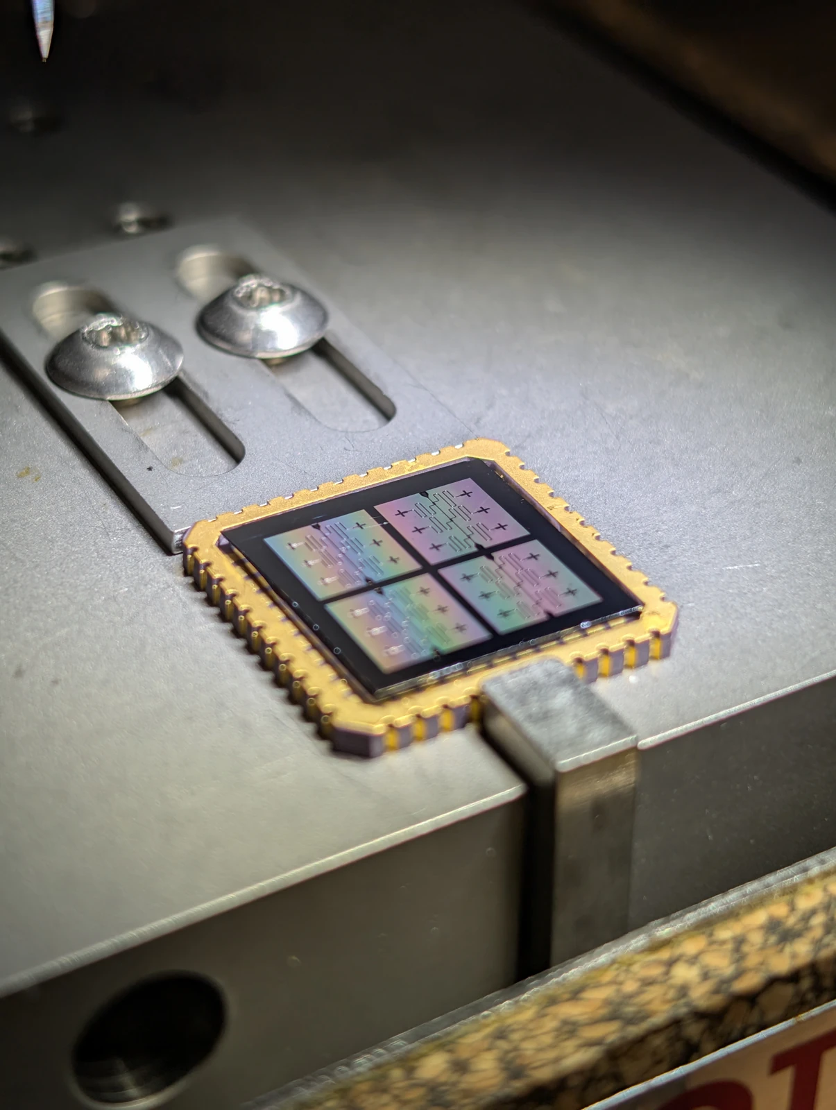 A small patterned chip sits in a gold-colored square package on a metal work surface beside two metal tools.