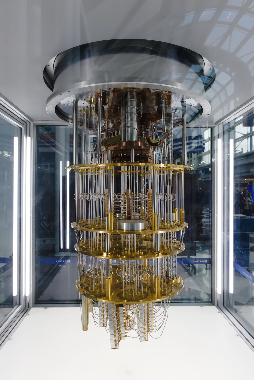 A gold-colored tiered cryogenic assembly hangs inside a cylindrical transparent enclosure in a bright airport interior.