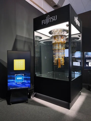 A Fujitsu-branded black exhibition stand surrounds a gold-colored assembly behind glass, with a display screen beside it.