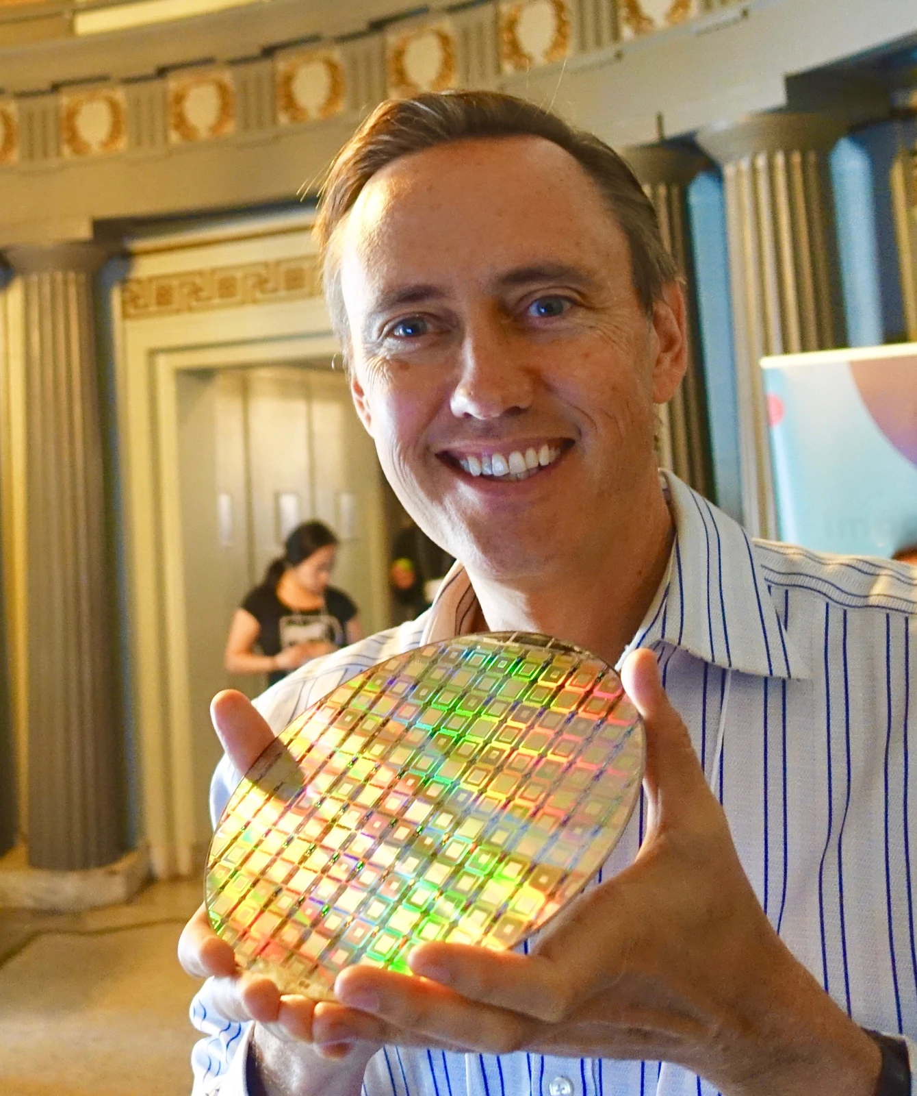 A smiling man holds a round iridescent wafer with a repeated grid pattern in front of him.