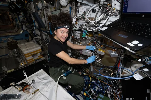 An astronaut wearing blue gloves handles cables and equipment inside a densely packed space-station workspace.