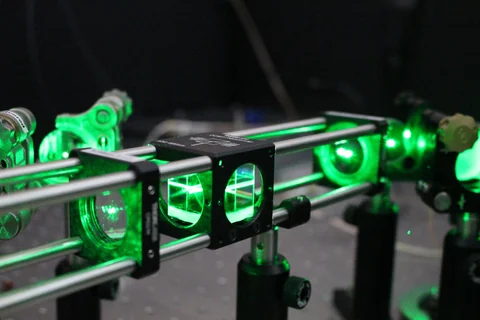 Bright green light passes through a row of small optical elements mounted on black posts against a dark background.
