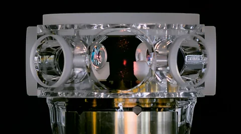 A transparent glass cell with circular windows surrounds a faint red spot above metallic vacuum hardware.