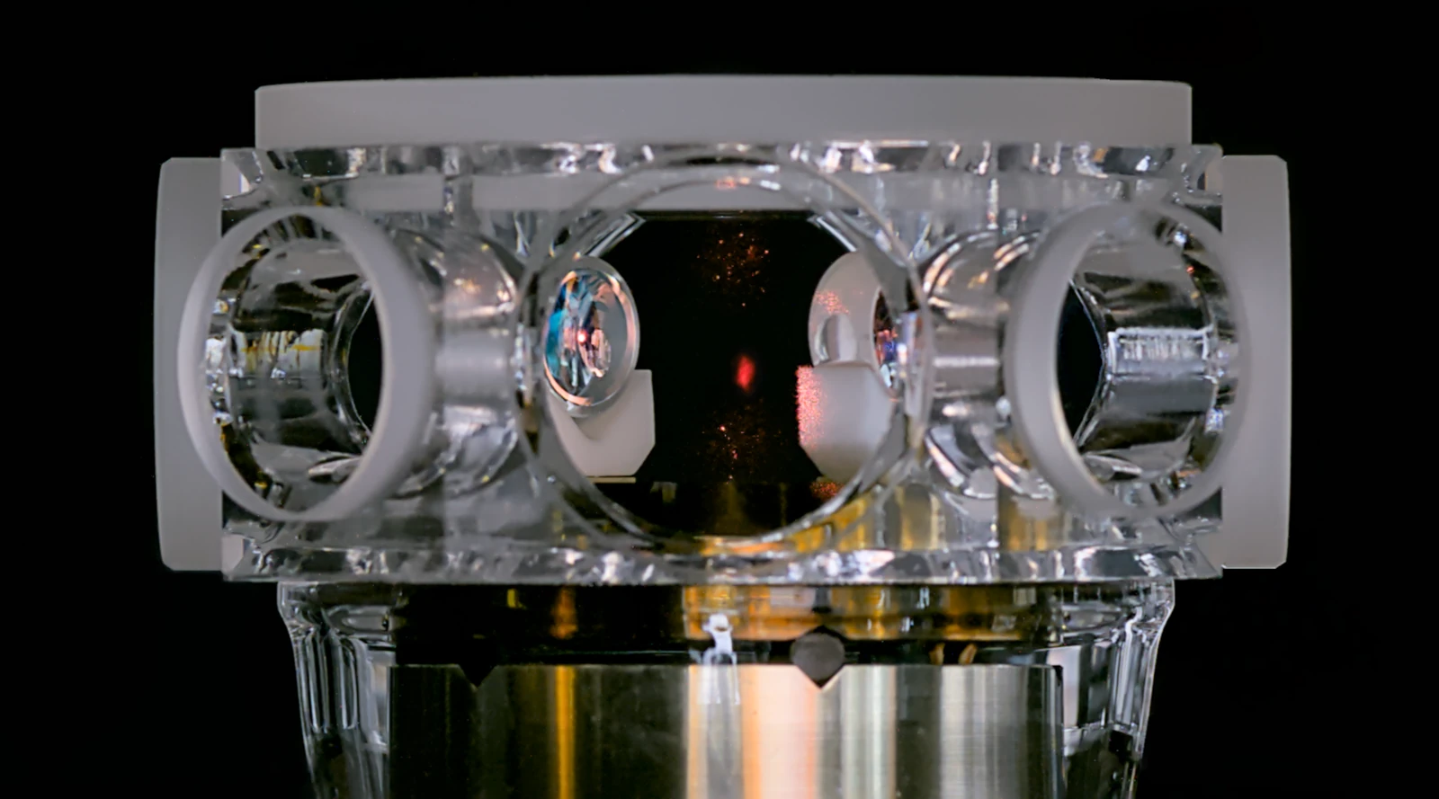 A transparent glass cell with circular windows surrounds a faint red spot above metallic vacuum hardware.