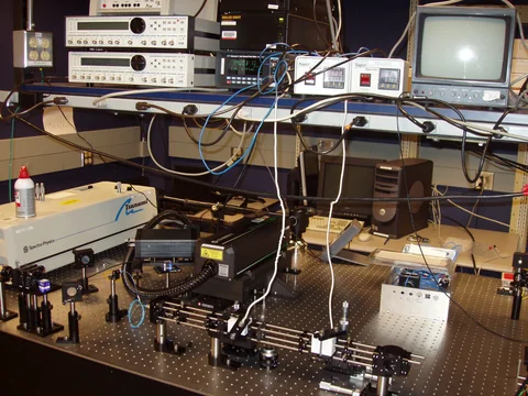Optical mounts and connected equipment cover a laboratory table beneath a row of electronic instruments and a monitor.