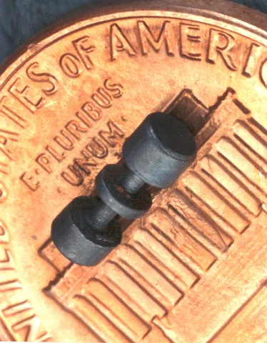 A small dark cylindrical metal component rests diagonally across a copper-colored U.S. penny.