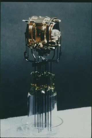 A small metal assembly with circular supports and fine pins stands against a dark blue background.