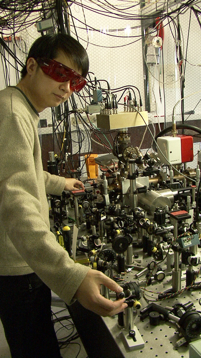 A researcher in protective glasses leans toward a dense optical table surrounded by cables and metal equipment.
