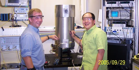 Two people wearing protective glasses stand beside a tall cylindrical instrument and racks of electronics.