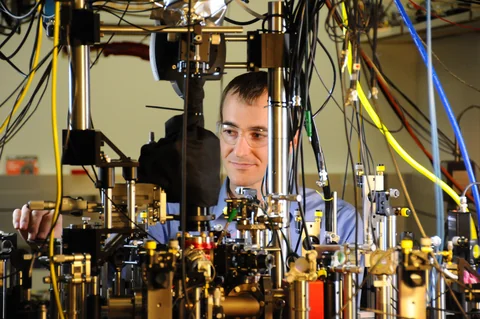 A researcher’s face is visible behind a crowded table of optical posts, components and hanging cables.