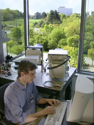 A man sits at a computer beside a telescope near a window looking toward another building and trees.