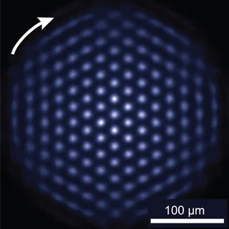 Blue-white points form a roughly circular triangular lattice against black, with a white arrow and a 100 micrometer scale bar.