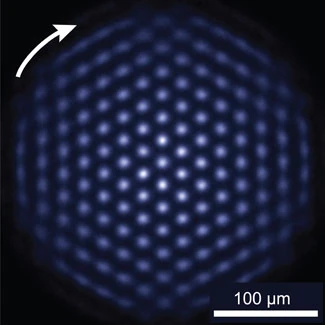 Blue-white points form a roughly circular triangular lattice against black, with a white arrow and a 100 micrometer scale bar.