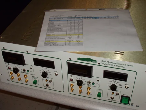 A silver electronic instrument has front-panel connectors, controls and a display, with a sheet of paper resting on top.