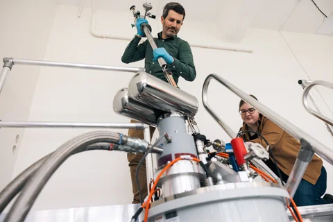 A person handles equipment above a large metal cylindrical apparatus while another person stands nearby.