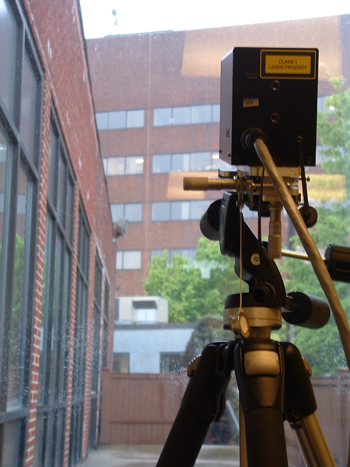 A black optical instrument on a tripod stands beside a window facing a brick building across an open space.