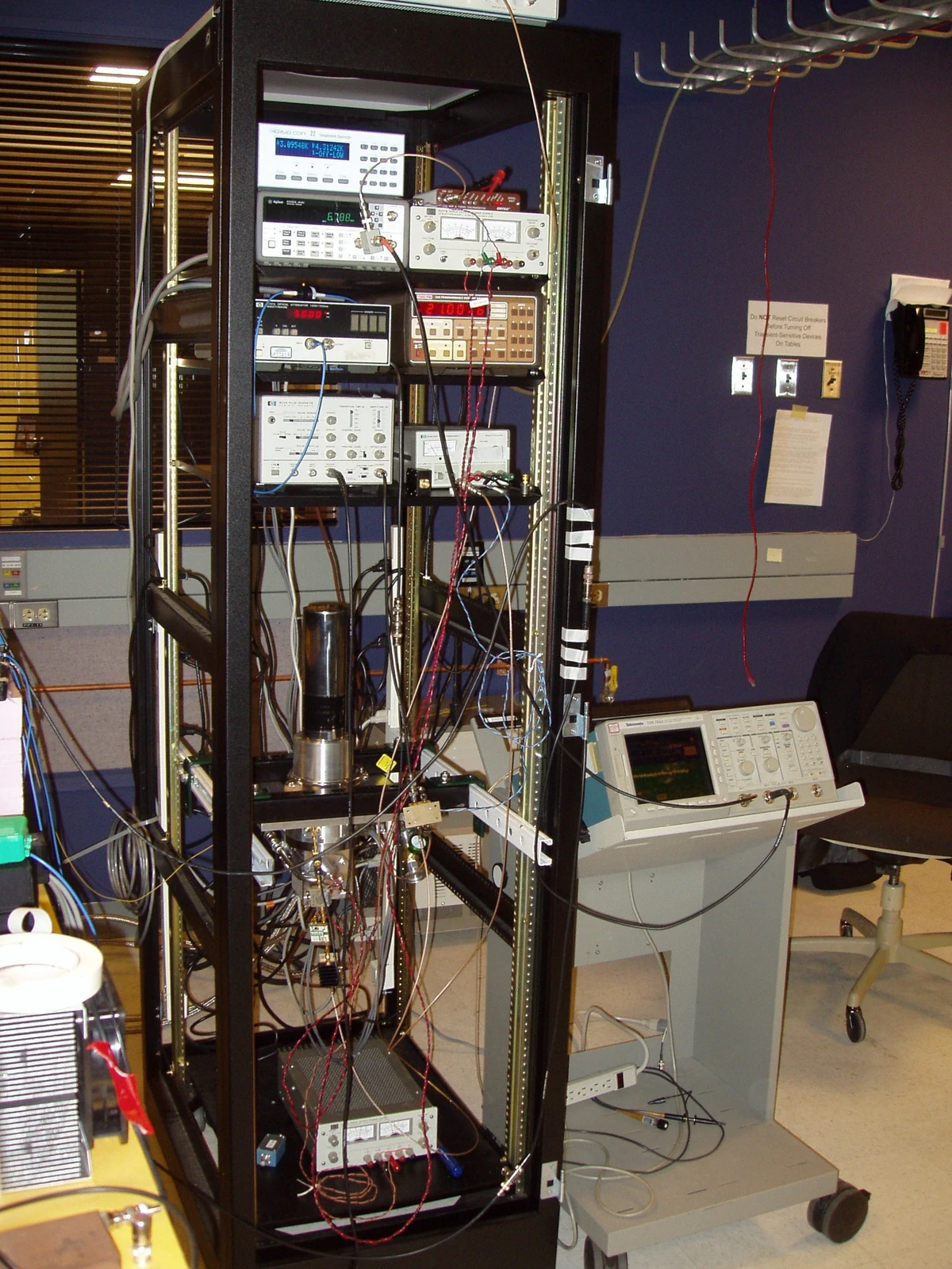 An open rack is filled with instruments, cables and a cylindrical component beside additional laboratory equipment.