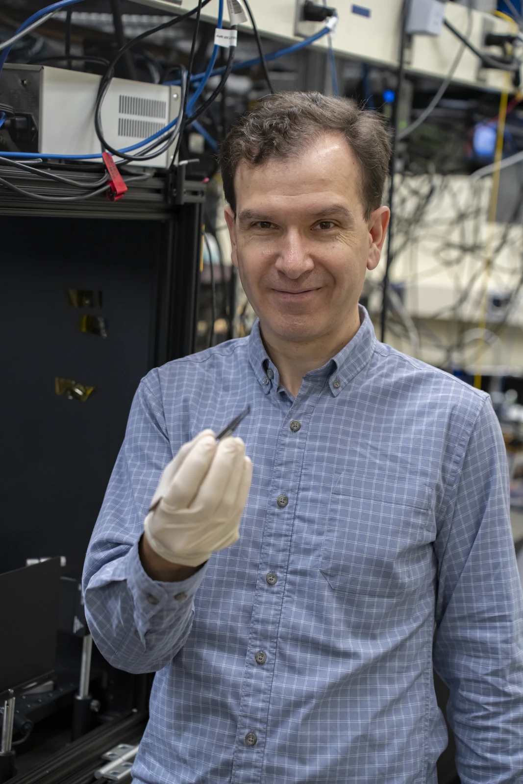 A researcher in a light blue checked shirt holds a small chip with tweezers in a white-gloved hand, with laboratory instruments behind him.