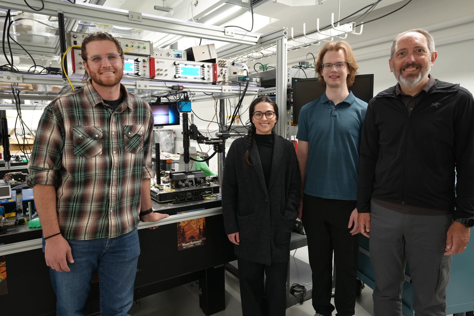 Four researchers stand beside an optical table and racks of instruments, cables and a display.