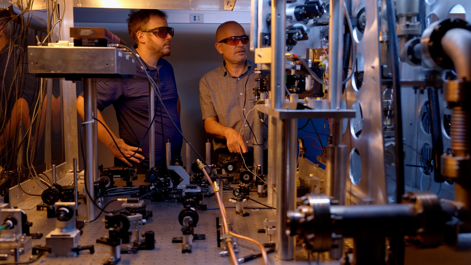 Two researchers wearing orange protective glasses lean toward metal optical assemblies and laboratory equipment.