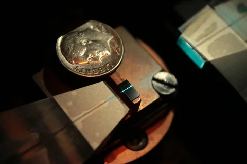 A dime sits above a small dark rectangular chip on a metal mount, with a bright blue line visible on the chip.