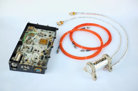 Open electronics enclosure at left, a coil of orange optical fibre in the centre, and a cylindrical metal sensor head at right on a pale surface.