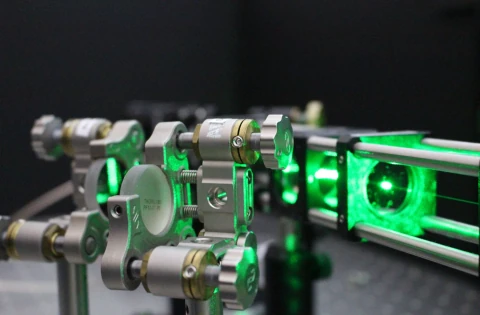 Green laser light illuminates a row of metal optical mounts and lenses on a dark laboratory table.