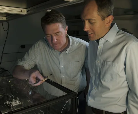 Two researchers inspect equipment together at an optical laboratory bench.