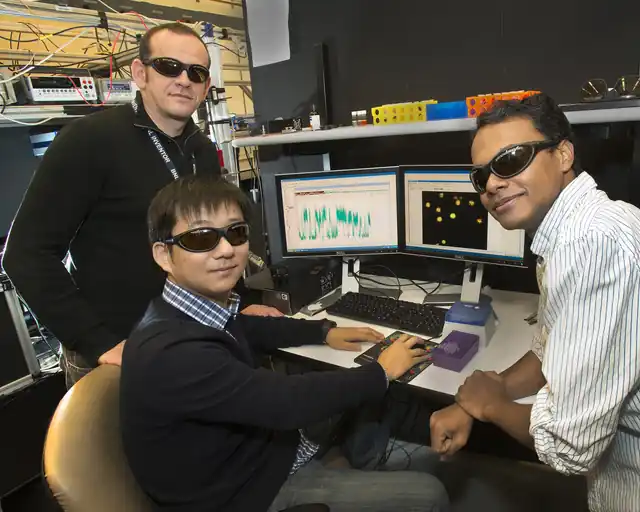 Archival context: Three researchers wearing protective glasses sit beside laboratory monitors and instruments.
