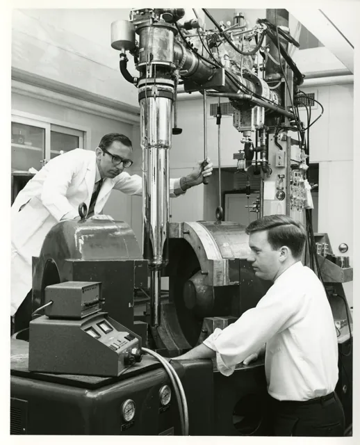 Two scientists position a tall Dewar between large magnet components in a black-and-white laboratory photograph.