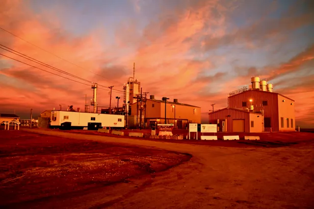 Industrial buildings and piping at a helium facility under an orange sunset sky.