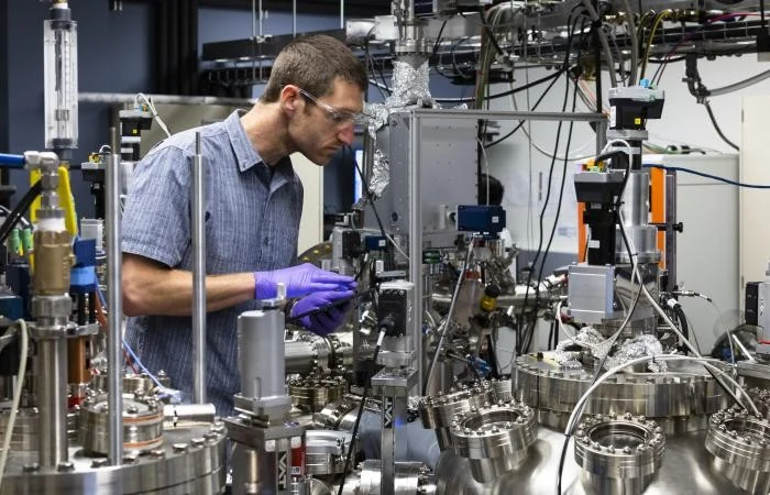 A researcher works beside a large metal vacuum apparatus in a materials laboratory.