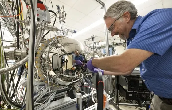 A researcher adjusts a large cylindrical instrument surrounded by cables and metal vacuum fittings.