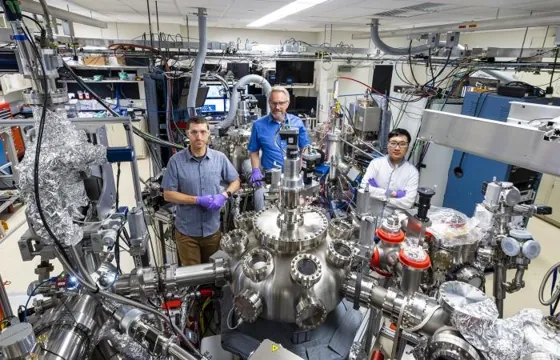Three researchers stand among metal vessels, instruments and cables in a materials laboratory.