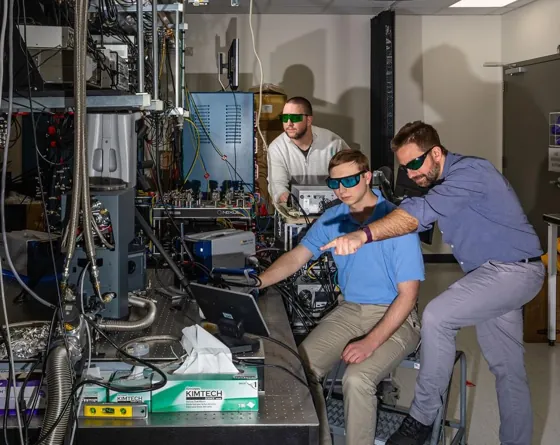 Three researchers wearing laser-safety glasses inspect a laboratory computer monitor.