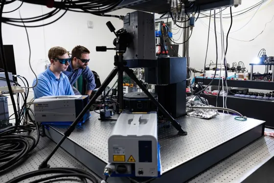 Two researchers work beside an ion-trap apparatus surrounded by vibrometer heads and optical equipment.