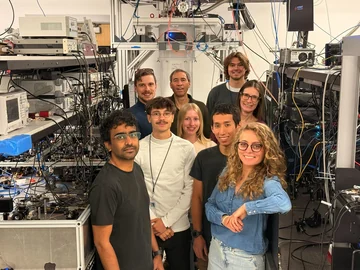 Nine team members standing between racks of laboratory electronics, optical equipment and cables.