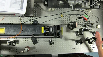 Top-down view of an optical table with a laser enclosure, mirrors, mounts, cables and a hand near the edge.